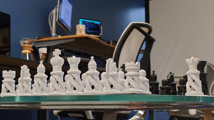 A 3D-printed chess set sits in the foreground while a software developer&rsquo;s station with two monitors, a coffee mug, and an office chair loom behind it.<br>
[photo credit: Ian Carroll]